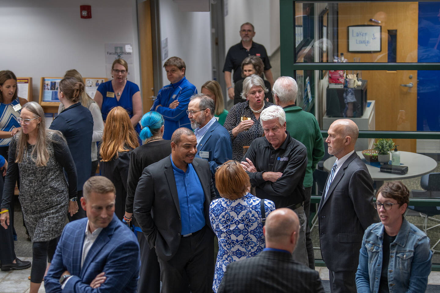 A diverse group of people engage in conversation in a professional indoor setting, with some standing near a glass-walled room and others gathered in a hallway.